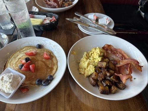 A plate of pancakes topped with strawberries and blueberries, alongside scrambled eggs, bacon, and seasoned potatoes.