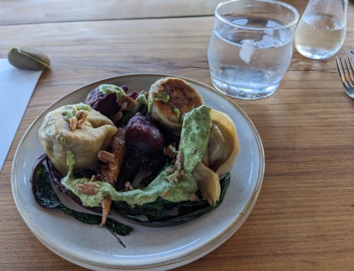 A plate of assorted dumplings and vegetables drizzled with green sauce, served on a wooden table with water glasses.