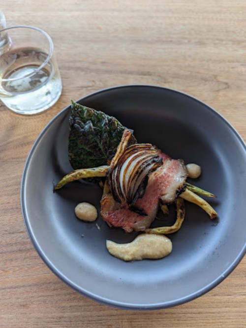 A plate of grilled meat with onions and greens, served with a creamy sauce, on a wooden table next to a glass of wine.