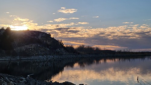 A serene landscape at sunset, with a calm lake reflecting the colorful sky and silhouetted trees along the shore.