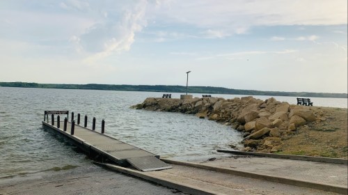 A calm lake scene featuring a wooden dock, rocky shore, and benches overlooking the water under a cloudy sky.