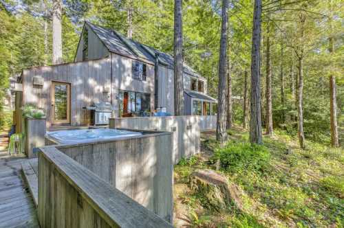 A modern wooden house nestled among tall trees, featuring a deck and hot tub in a serene forest setting.