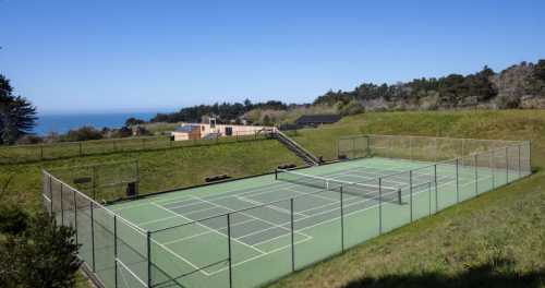 A green tennis court surrounded by grass, with a building and ocean view in the background under a clear blue sky.