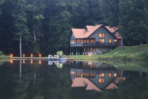 A serene lakeside house with warm lights reflecting on the water, surrounded by trees at dusk.