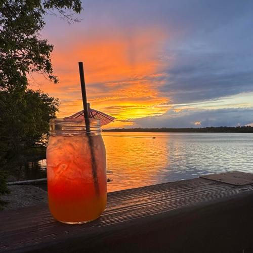 A colorful sunset over water, with a cocktail in a jar featuring a small umbrella on a wooden railing.