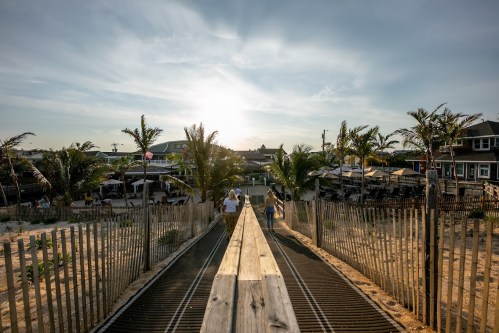 A wooden walkway leads to a beach at sunset, flanked by palm trees and beachside buildings.