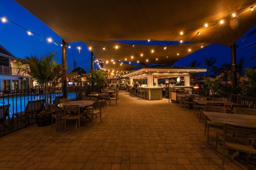 A cozy outdoor dining area with string lights, tables, and a bar near a pool, set against a twilight sky.