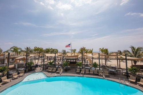 A pool area with lounge chairs, palm trees, and an American flag, overlooking a sandy beach under a blue sky.