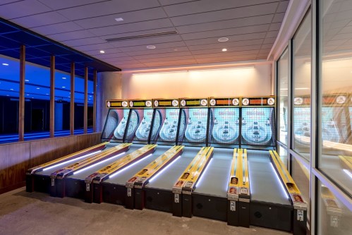 A row of illuminated skee-ball machines in a modern game room with blue lighting and a sleek design.