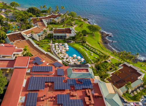 Aerial view of a resort with solar panels, a pool, and palm trees by the ocean.