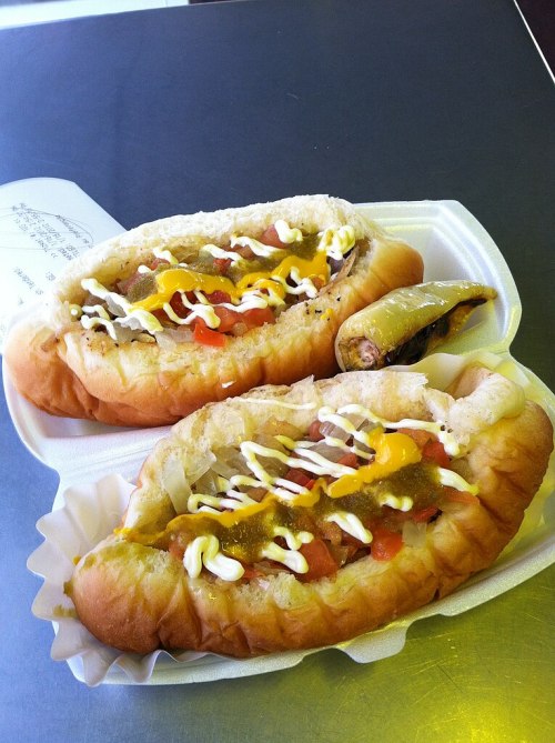 Two hot dogs in buns topped with mustard, mayonnaise, diced tomatoes, and a side of pickled pepper on a white tray.