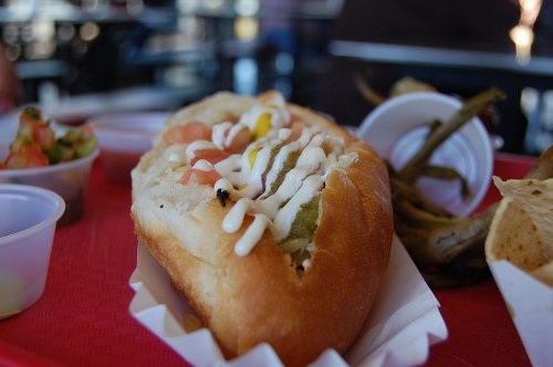 A close-up of a hot dog topped with mustard, mayonnaise, and diced tomatoes, served on a red tray with sides.