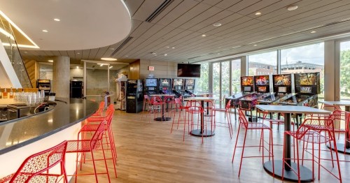Modern lounge area with red chairs, tables, and vending machines, featuring large windows and a sleek bar.