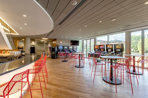 Modern lounge area with red chairs, tables, and arcade machines, featuring large windows and a sleek bar.