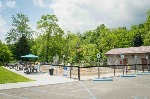 A fenced playground area with picnic tables, a splash pad, and restrooms, surrounded by trees under a cloudy sky.