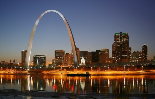 The Gateway Arch illuminated at dusk, reflecting in the water with a city skyline in the background.