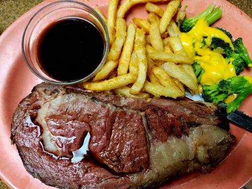 A plate featuring a large slice of roast beef, fries, broccoli with cheese, and a small bowl of sauce.