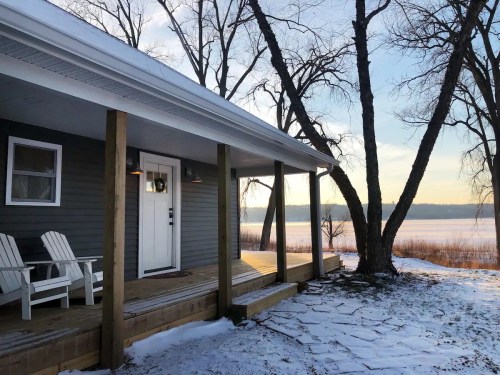 A cozy house with a porch overlooking a frozen lake, surrounded by trees and snow.