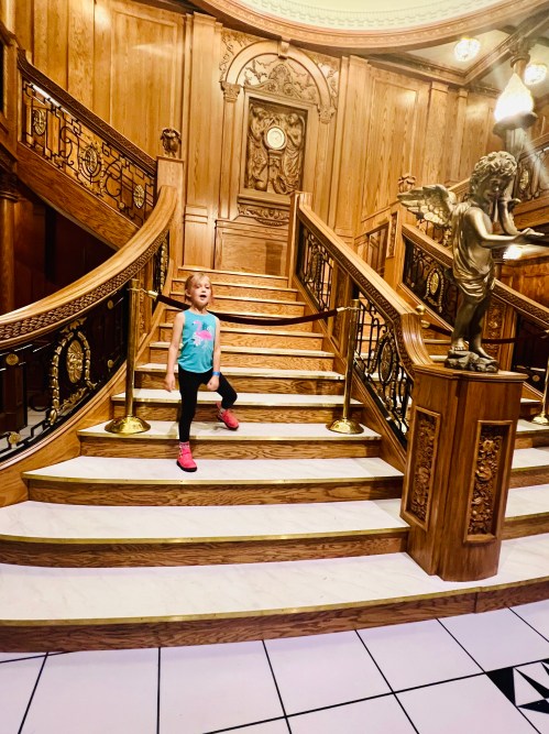 A young girl stands on a grand wooden staircase with ornate railings and a statue, smiling in a brightly lit space.
