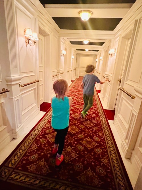 Two children run down a long, elegant hallway with white walls and a red carpet, illuminated by wall sconces.