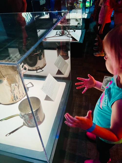 A child gazes at a glass display case containing a pot, with other visitors in the background.