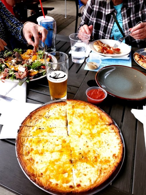 A table with a large cheese pizza, a salad, a drink, and plates of food, set in a casual dining environment.