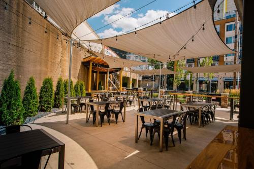 Outdoor dining area with tables and chairs under a large canopy, surrounded by greenery and modern buildings.