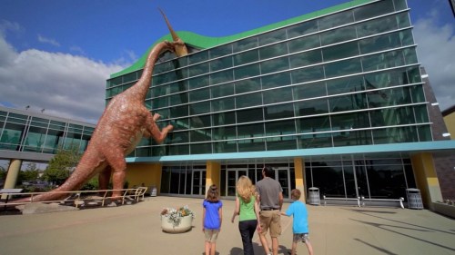 A family approaches a building with a large dinosaur sculpture on the roof, under a blue sky.