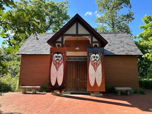 A brick building with a sloped roof, featuring large cicada banners on either side of the entrance. Green trees surround it.