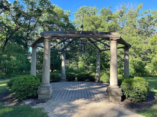 A stone gazebo with four columns surrounded by lush greenery and a paved pathway. Clear blue sky above.