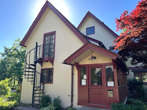 A charming two-story house with a spiral staircase, surrounded by greenery and a vibrant red tree under a clear blue sky.