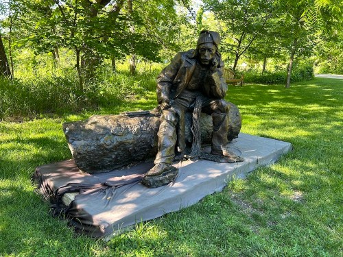 A bronze statue of a seated figure in a hat, resting on a log, surrounded by greenery in a park.