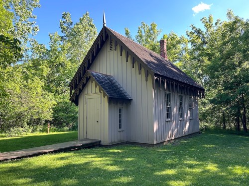 A small, charming wooden building with a peaked roof, surrounded by lush greenery and a clear blue sky.
