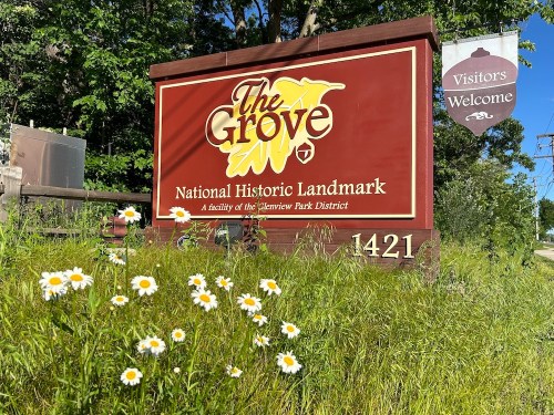 Sign for The Grove, a National Historic Landmark, surrounded by greenery and wildflowers, with a "Visitors Welcome" banner.