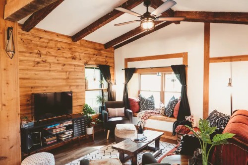 Cozy living room with wooden beams, a TV, seating area, and a window nook filled with cushions and plants.