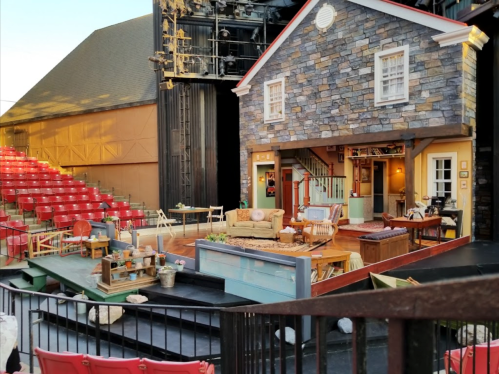 A theater stage set designed as a cozy house, featuring furniture and decor, with red audience seats in the background.