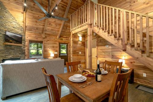 Cozy wooden cabin interior with a dining table, staircase, and living area featuring a stone wall and large windows.