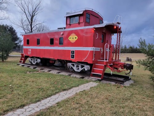 A red caboose on a track, set in a grassy area with trees and a cloudy sky in the background.