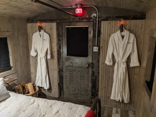 Cozy interior of a cabin with two white robes hanging, a bed, and an exit sign above a metal door.