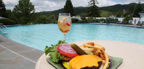 A cheeseburger with lettuce, tomato, and fries beside a fruity drink, set against a scenic pool and mountain backdrop.