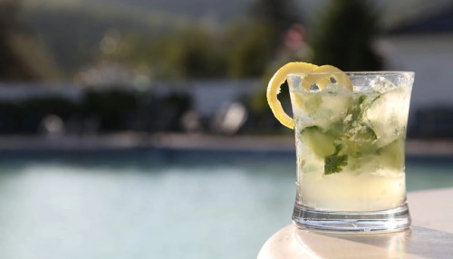 A refreshing cocktail with mint and lemon slices sits on a table by a pool, with a blurred background of greenery.