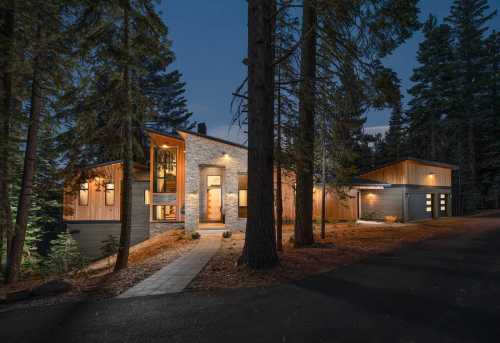 A modern house surrounded by tall trees, illuminated at dusk with a pathway leading to the entrance.