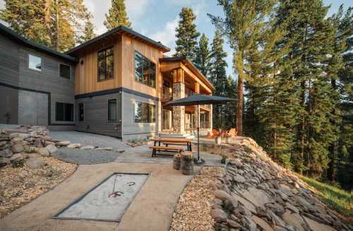 Modern wooden house surrounded by tall trees, featuring a patio with seating and a gravel pathway.