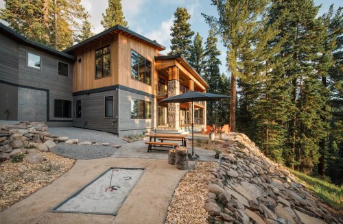 Modern wooden house surrounded by tall trees, featuring a patio with seating and a gravel pathway.