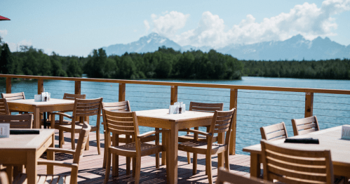 Outdoor dining area with wooden tables overlooking a lake and mountains under a clear blue sky.