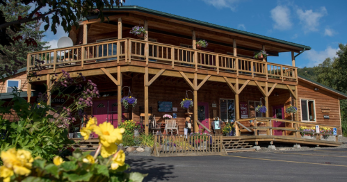 A wooden building with a balcony, surrounded by flowers and greenery, under a blue sky with scattered clouds.