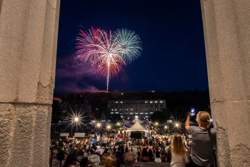 A crowd watches colorful fireworks explode in the night sky, framed by stone columns.