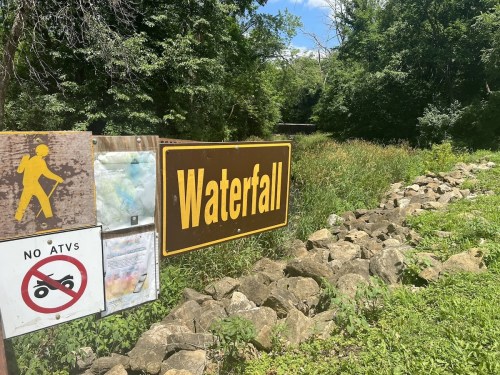A sign indicating a waterfall, with a hiker symbol and a "No ATVs" sign, surrounded by greenery and rocks.