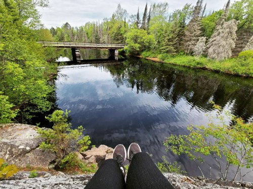 A person sits on a rock by a calm river, with a bridge and lush greenery in the background.