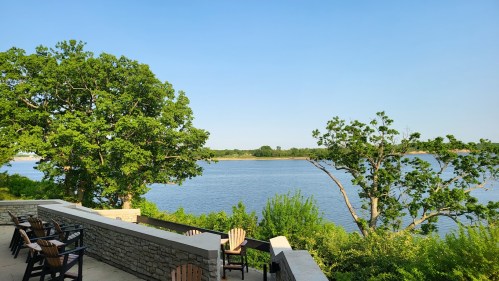 A serene river view with lush green trees and a stone patio featuring wooden chairs under a clear blue sky.